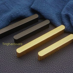 Op de afbeelding: Een verzameling rechthoekige kastgrepen in verschillende afwerkingen. De grepen zijn in zwart, donker brons en goud. Ze zijn gerangschikt op een donkere ondergrond met een blauwe stof op de achtergrond. De tekst "Tinghardware" is zichtbaar.