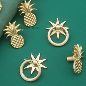 以下が含まれることがあります： パイナップルとスターバーストのデザインが施された、ゴールドカラーのキャビネットハードウェア。 パイナップル型のノブとスターバーストリングプル、ステム付きのパイナップル型プルが含まれています。 ハードウェアは緑色の表面に表示されています。