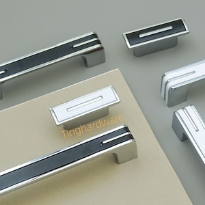 May include: A collection of cabinet hardware, including handles and knobs. The handles have a rectangular design with a black or white center stripe and chrome accents. The knobs are rectangular with a single stripe.