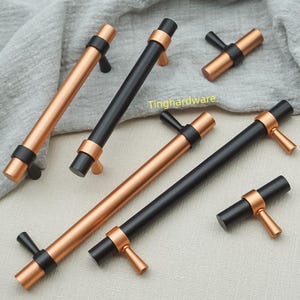 May include: Cabinet handles in copper and black. The cylindrical handles have a T-shaped pull. The hardware is displayed on a neutral-coloured fabric. The image includes the text "Tinghardware".