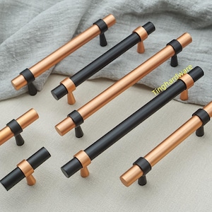 May include: A collection of cabinet pulls in various sizes. The pulls feature a combination of black and copper-toned metal. The handles have a cylindrical design with a black center section and copper-toned ends. The pulls are arranged on a neutral-colored fabric.