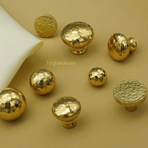 May include: A collection of gold-colored cabinet knobs and pulls with a hammered texture. The hardware pieces vary in size and shape, including round and mushroom-style designs. The knobs are arranged on a light brown surface.