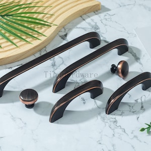 Op de afbeelding: Een collectie bronzen kastbeslag, waaronder handgrepen en knoppen. De handgrepen zijn gebogen en verkrijgbaar in verschillende maten. De knoppen zijn rond. Het beslag heeft een donkerbronzen afwerking met koperen accenten.