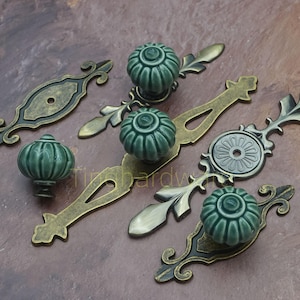 Może przedstawiać: Zestaw okuć do szafek w stylu antycznym. Zestaw zawiera zielone ceramiczne gałki i dekoracyjne płytki tylne w odcieniach brązu i mosiądzu. Gałki mają żebrowany wzór, a płytki tylne zdobione są ornamentami kwiatowymi i zwijanymi.
