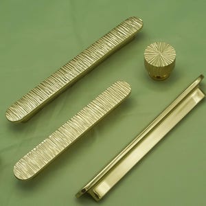 May include: A collection of gold-colored cabinet hardware, including two long bar pulls with textured surfaces, a round knob with a radial pattern, and a sleek, flat pull. The hardware is arranged on a green surface.