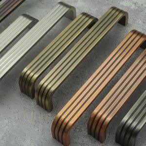 May include: An assortment of cabinet handles in different metallic shades, such as silver, bronze, and copper. Each handle features a curved, arched shape with several parallel grooves, providing a contemporary look. The hardware is displayed on a neutral grey surface.