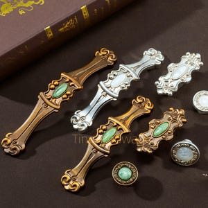May include: An assortment of decorative cabinet hardware, including bronze, silver, and gold-toned handles and knobs. The handles feature ornate designs with oval-shaped, green and white accents. The knobs have a circular design with a central stone.