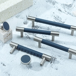 May include: A collection of cabinet hardware, including handles and knobs. The handles have a brushed silver finish with a navy blue leather-wrapped center. The knobs are round with a matching navy blue center. The hardware is displayed on a light blue surface.