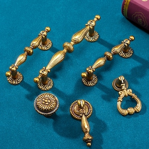 Peut inclure: Ensemble de quincaillerie de meubles en laiton antique, comprenant des poignées et des boutons. Le matériel présente des détails ornés et une finition dorée chaleureuse. Les pièces sont disposées sur une surface turquoise, mettant en valeur leurs motifs.