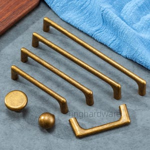 以下が含まれることがあります： アンティーク真鍮製のキャビネット金具のコレクション。4つの異なるサイズのバープル、丸いノブ、球形のノブ、U字型のハンドルが含まれています。金具はグレーの表面に配置されています。