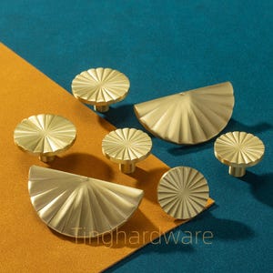 Może przedstawiać: Kolekcja złotych gałek i uchwytów do szafek. Gałki są okrągłe z wzorem promieni słonecznych, a uchwyty mają kształt wachlarza. Obraz jest umieszczony na tle w kolorze turkusowym i musztardowym. Widoczny jest tekst "Tinghardware".