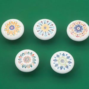 May include: Five white ceramic cabinet knobs with colourful floral designs. The knobs feature orange, blue, yellow, and green patterns. The knobs are arranged on a solid green background.