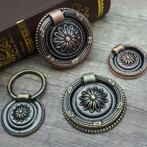 May include: A collection of antique-style cabinet hardware, including drawer pulls and knobs. The hardware features a floral design in bronze, copper, and brass finishes. The pieces vary in size and style, with ring pulls and round knobs.