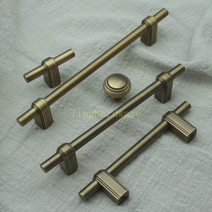 May include: An assortment of antique brass cabinet hardware, including handles and a knob. The handles vary in length, with a cylindrical bar and rectangular end pieces. The knob is round with a slightly domed top. The hardware is displayed on a neutral fabric.