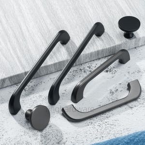 May include: A collection of matte black and gray cabinet hardware, including handles and knobs. The handles are curved and come in various sizes. The knobs are round and have a simple design. The hardware is displayed on a light gray surface.