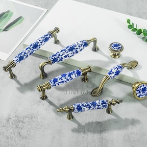 May include: A collection of cabinet hardware featuring white ceramic handles with blue floral patterns. The handles have brass-colored metal bases and accents. Includes knobs and pulls in various sizes and shapes.