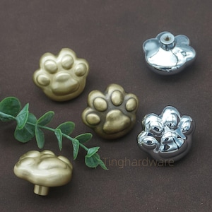 Puede incluir: Un conjunto de pomos para armarios con forma de pata, en acabados metálicos dorados y plateados. Los pomos se presentan sobre una superficie marrón con una ramita de vegetación, mostrando su diseño decorativo.