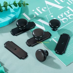 Op de afbeelding: Een assortiment zwarte en bronzen kastknoppen en -grepen. De knoppen zijn rond en de grepen zijn rechthoekig met decoratieve randen. De hardware wordt gepresenteerd op een lichtblauwe achtergrond.