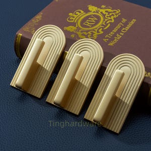 Könnte beinhalten: Drei goldfarbene Schrankgriffe mit rechteckiger Basis und gebogenem, bogenförmigem Design. Jeder Griff hat einen vertikalen Stab zum Greifen. Die Griffe sind auf einer dunkelblauen Oberfläche angeordnet, mit einem Buch im Hintergrund.