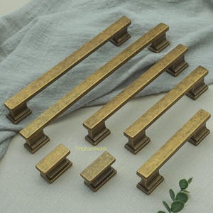 May include: A selection of antique brass cabinet pulls and knobs. The pulls are rectangular with a raised centre and square bases. The knobs are smaller, rectangular, and match the pulls. The hardware is arranged on a grey fabric.