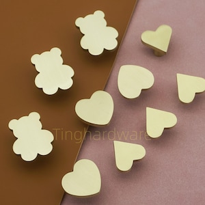 Puede incluir: Una colección de tiradores de armario de latón con forma de osos de peluche y corazones. Los tiradores son de varios tamaños y se muestran sobre un fondo marrón y rosa. El texto "Tinghardware" es visible.