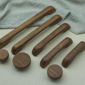 May include: A selection of wooden cabinet handles and knobs in a warm brown hue. The image shows several long, curved handles and round knobs, all displaying visible wood grain. The longest handle is approximately 30 cm in length.