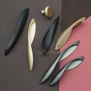 以下が含まれることがあります： 黒、金、銀色の取っ手と丸いノブを含む、モダンなキャビネットハードウェアのコレクション。 ハンドルは湾曲した葉のようなデザインで、ノブはシンプルで丸みを帯びた形状です。 ハードウェアは、ツートンカラーの茶色とピンクの背景に表示されます。