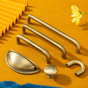 Peut inclure: Un assortiment de quincaillerie d'armoire en laiton antique, comprenant trois poignées de barre, une poignée de tasse, un bouton rond et une poignée incurvée. L'ensemble est présenté sur une surface jaune avec un tissu bleu et une fleur jaune.