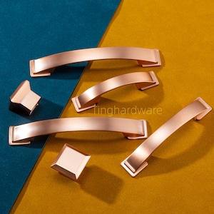 以下が含まれることがあります： ローズゴールドのキャビネットハードウェアセット。カーブしたハンドルと正方形のノブが含まれています。ハードウェアは、ターコイズとゴールドの背景に表示されます。「tinghardware」の文字が背景に表示されます。