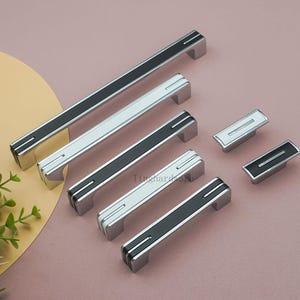 May include: A collection of silver cabinet pulls with black and white accents. The pulls are rectangular with a central stripe and two smaller accent lines. The collection includes both long bar pulls and smaller knob-style pulls.