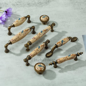 May include: A selection of antique-style cabinet handles and knobs. The cream-coloured ceramic surfaces are decorated with a bird and floral design, set on a bronze-toned metal base. The text "Tinghardware" is visible. Ideal for furniture restoration.