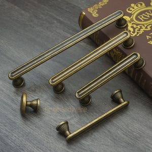 Puede incluir: Un surtido de herrajes de gabinete de latón antiguo, que incluyen manijas y un pomo. Las manijas varían en tamaño, con un diseño acanalado y piezas decorativas en los extremos. El hardware se muestra sobre una superficie oscura con vetas de madera.