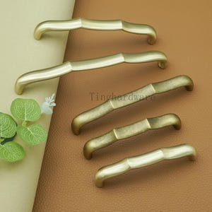 May include: A collection of five cabinet pulls in varying sizes and finishes. The pulls have a bamboo-inspired design, with a curved handle and a textured center. The finishes range from a bright gold to a more antique brass. The text "Tinghardware" is visible.