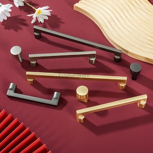 May include: A collection of cabinet hardware, including handles and knobs, in various metallic finishes such as gold, silver, and dark gray. The hardware is displayed on a burgundy surface with a wooden accent and floral decorations.