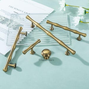 May include: A selection of antique brass cabinet handles and a knob, displayed on a clear glass surface. The hardware features a bamboo-style design with a warm, aged finish. The text "Tinghardware" is visible.
