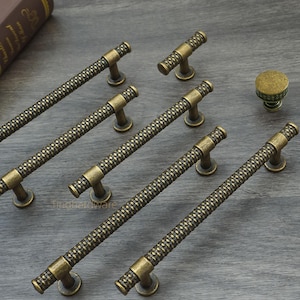 Könnte beinhalten: Antik-Messing-Schrankbeschlagsatz. Das Set enthält mehrere lange Stangengriffe und kleinere Knöpfe. Die Griffe haben ein strukturiertes, gewebtes Muster, und die Knöpfe haben ein dekoratives Design. Die Hardware wird auf einer grauen Holzoberfläche präsentiert.