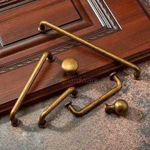 Op de afbeelding: Een assortiment antiek messing kastbeslag, inclusief handgrepen en knoppen, gepresenteerd tegen een donkere houten achtergrond. Het beslag heeft een vintage uitstraling, met zichtbare slijtage. De tekst "Tinghardware" is zichtbaar.