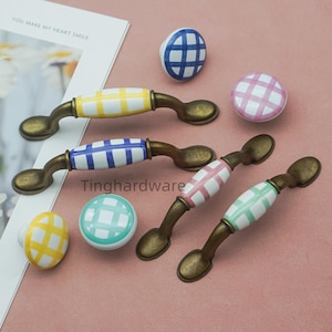 May include: A collection of colorful cabinet hardware, including drawer pulls and knobs. The pulls have bronze-colored metal bases with ceramic handles in yellow, blue, pink, and green, each with a grid pattern. Knobs in matching colors and patterns are also present.