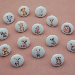 May include: A collection of white ceramic cabinet knobs, each featuring a different animal illustration. Animals include a lion cub, elephant, kitten, giraffe, teddy bear, dinosaur, hedgehog, zebra, puppy, monkey, rabbit, lamb, wolf, fox, and tiger cub.