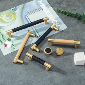 Peut inclure: Ensemble de quincaillerie d'armoires avec des détails en laiton. Les poignées ont des poignées noires et claires en bois. Deux boutons sont également inclus. La quincaillerie est présentée sur une surface grise avec un magazine en arrière-plan.