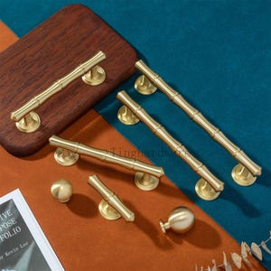 Pode incluir: Uma coleção de ferragens para armários em tom de latão, incluindo puxadores em estilo bambu e puxadores redondos. As alças variam em tamanho, algumas colocadas sobre uma superfície de madeira. Fundo azul, castanho e laranja.