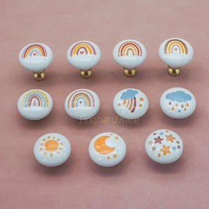 Pode incluir: Uma coleção de doze puxadores de armário de cerâmica branca com bases de latão. Cada puxador apresenta um design colorido diferente, incluindo arco-íris, nuvens, um sol, uma lua crescente e estrelas. Os puxadores estão dispostos sobre uma superfície rosa.