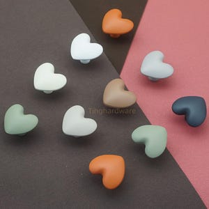 May include: A collection of heart-shaped cabinet knobs in various colors, including white, orange, green, and gray. The knobs are arranged on a brown and pink background. The brand name "Tinghardware" is visible.