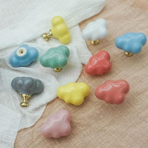 May include: A selection of cloud-shaped cabinet knobs in pastel shades such as yellow, blue, pink, and green. Each knob has a brass base. The knobs are displayed on a white cloth and a beige surface.