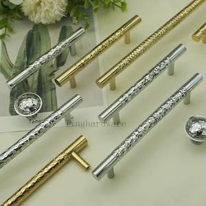 Op de afbeelding: Een assortiment kastgrepen en knoppen in zilver- en goudtinten. De handgrepen hebben een getextureerd, gehamerd patroon, terwijl de knoppen een facetontwerp hebben. De tekst "Tinghardware" is zichtbaar op het oppervlak.