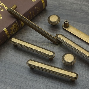 Może przedstawiać: Asortyment antycznych mosiężnych okuć do szafek, w tym uchwytów i gałek. Uchwyty mają prostokątny kształt z teksturowanym wzorem rombu i gładką, owalną sekcją środkową. Gałki są okrągłe.