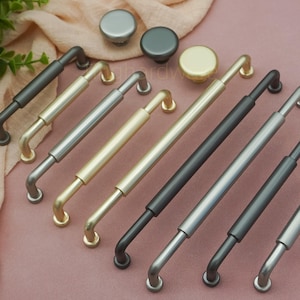 May include: A collection of cabinet hardware, including various pull handles and knobs. The handles come in several metallic finishes, such as gold, silver, and black. The knobs are round and match the handle finishes. The hardware is arranged on a pink surface.