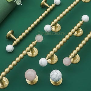 May include: Gold-toned cabinet pulls with marble and pink quartz accents. The pulls have a beaded design and are arranged on a green surface. The text "Tinghardware" is visible.