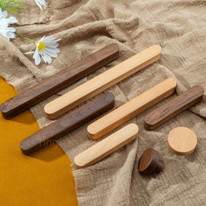 May include: A selection of wooden cabinet pulls and knobs in different shapes and sizes. The pulls are rectangular, and the knobs are round, with a mix of light and dark wood tones. The items are displayed on a neutral-coloured fabric.
