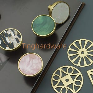 Puede incluir: Un conjunto de tiradores decorativos para armarios y recortes de latón. Los tiradores presentan diseños circulares en negro, verde, blanco y rosa, con bases doradas. Los recortes de latón incluyen formas circulares y cuadradas.